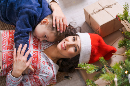 Happy mom with son in santa hat lie on floor at home under tree with gifts, enjoy christmas and new year holidays. Joyful family on christmas eveの写真素材