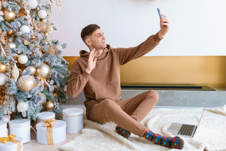 Young caucasian man communicates via video communication using phone in living room while sitting on the floor near the Christmas tree. Happy New Year wishes to relatives and friends onlineの写真素材