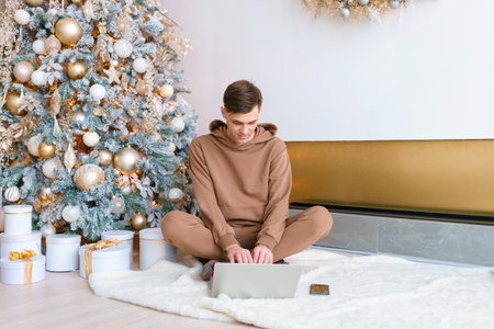 A man sitting on floor near Christmas tree orders online goods on black friday using a laptop and phone. Happy guy in cozy clothes communicates in social networksの写真素材