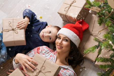 Top view mother and son in santa hats looking at camera and smiling while lying on floor at home near new year tree with gifts, enjoy winter holidaysの写真素材