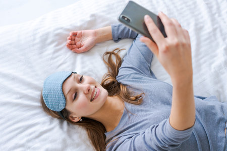 Beautiful woman in blue pajamas looking into phone. Morning starts with phone. Girl in sleep mask in bright spacious room. Reading news from phone. Attractive girl holding a phone and looking at itの写真素材
