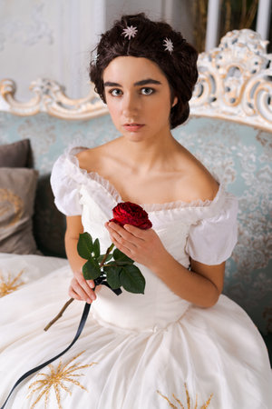 Portrait young beautiful attractive bride with red rose in her hand. Vintage white dress and bridal jewelry. Sitting on the couchの写真素材
