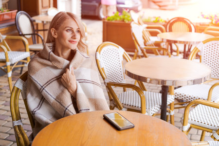 Young woman while relaxing in cafe at table on street, happy caucasian woman is wrapped in a warm cozy plaid, when she drinks coffee in a cafe in her free time, the phone lies on the table.の写真素材