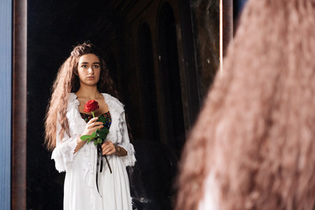 Attractive woman with long brown hair holds luxurious red rose. She is wearing a white vintage dress looking into the reflection in the mirror. Caucasian girl in a romantic moodの写真素材