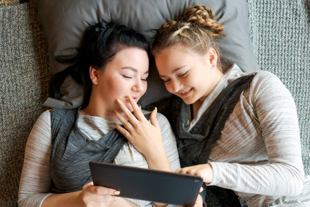 Happy two sisters using tablet while lying on their backs on floor at home laughing cheerfully watching videos or chatting online, wearing gray clothesの写真素材