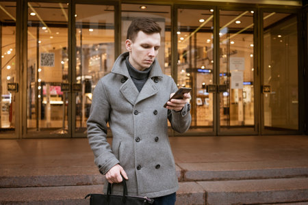 Smiling young man in gray coat is holding scroll text messages in his mobile phone on street in city...