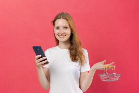 Beautiful caucasian young woman holding small shopping cart and mobile phone in white t-shirt for mockup isolated on pink background, online shopping conceptの写真素材