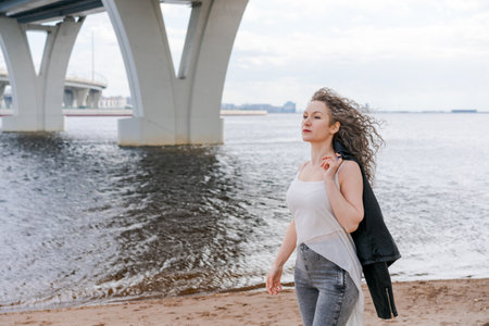 Young stylish woman in light light T-shirt and leather jacket and curly hair flying along river embankment is blowing in wind. Summer holidays, walk by the river in the afternoon on a day offの写真素材
