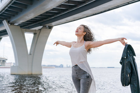 Attractive woman enjoys summer walks along the promenade. Wears T-shirt and leather jacket, her hair blows in the wind in cloudy weather. Summer vacation concept.の写真素材