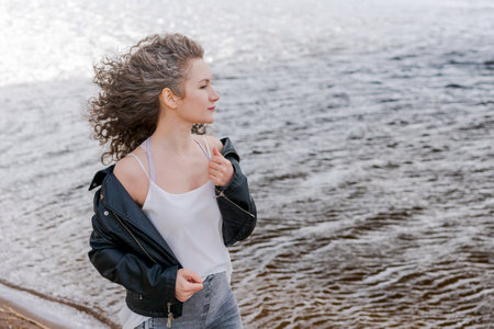 Young stylish woman in light light T-shirt and leather jacket and curly hair flying along river embankment is blowing in wind. Summer holidays, walk by the river in the afternoon on a day offの写真素材