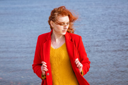 Attractive redhead woman walks on street in city near river, weekend outdoors, the woman wears a red coat and a yellow sweater, enjoys a warm vertの写真素材
