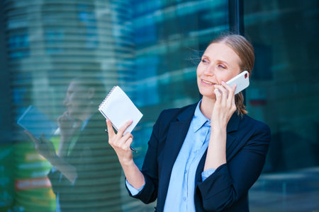 Smart professional caucasian business woman uses mobile phone in city outdoors holding documents in her hands, dressed in a blue shirt on the background of a business centerの写真素材