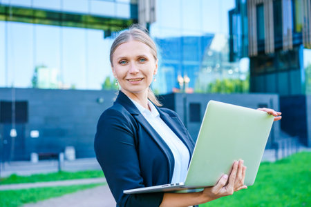 Portrait confident successful young woman, formally dressed, business woman, standing with a laptop outdoors against the backdrop of a business centerの写真素材