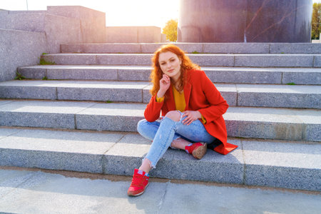 Young redhead woman in red coat yellow sweater and jeans sits on stairs in afternoon in sunny weather in the park in springの写真素材