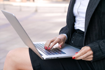 Young serious business woman with laptop studying online while sitting on bench against office center background, business woman working on laptop pensive and focusedの写真素材
