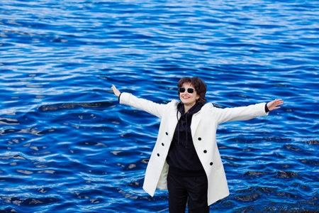 Cool hipster young girl in black hoodie and white coat with short haircut in sunglasses standing by the river in the city enjoying the sun in spring gesturing with her hands in happinessの写真素材