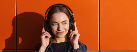 Portrait an attractive young woman smiling with wireless headphones against an orange wall in casual...