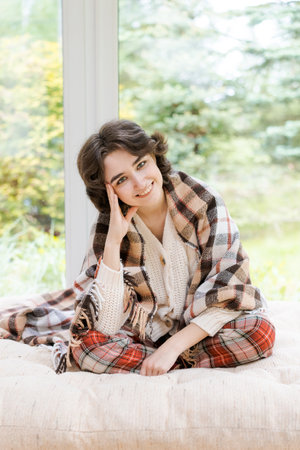 Portrait lonely caucasian young woman sitting near window apartments in a white sweater and a red plaid, spends time in the morning on the veranda in a country houseの写真素材