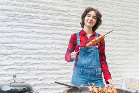 Barbeque party in garden with young woman in denim overalls and red plaid shirt in a country house on the terrace preparing meat skewersの写真素材
