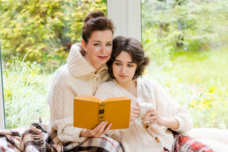 Nice girl and her mother enjoy sunny morning. Good time at home they sit on veranda against background windows with blanket and mug morning coffee concept relationship between children and parentsの写真素材