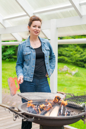 Woman cooks barbecue for her family outside in garden in country house wearing casual clothes and a denim jacketの写真素材