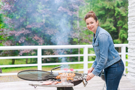 Woman cooks barbecue for her family outside in garden in country house wearing casual clothes and a denim jacketの写真素材