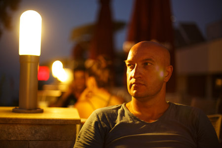 a bald man sits in the evening in a street cafe in the light of lanterns and looks to the side,...