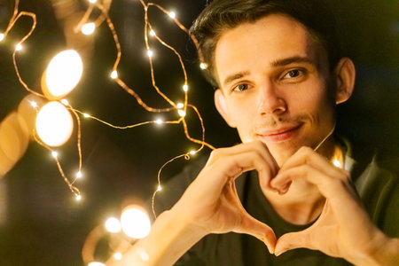a young guy at night holding a glowing garland shows a heart gesture with his hands, selective focusの写真素材