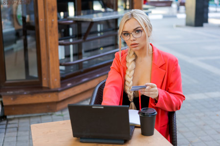 Beautiful woman sits at table in street cafe and works on tablet. Concept remote work, free work schedule. In a bright pink jacket and glassesの写真素材