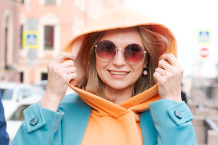 Happy woman walking through city with bag in her hand, dressed in bright clothes, an orange hoodie and a blue coat, smiling contentedly in sunglassesの写真素材