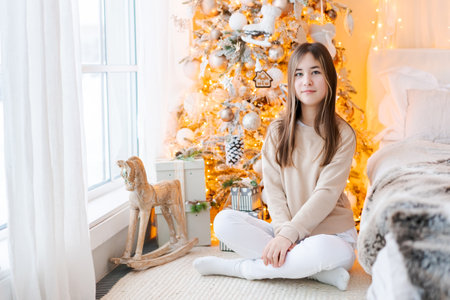 Cute girl sits on floor by window with Christmas tree in background in her bedroomの写真素材