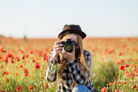 A woman wearing hat and glasses is taking picture field red flowers. Concept of serenity and beauty, as woman captures the vibrant colors flowers in her photographの写真素材
