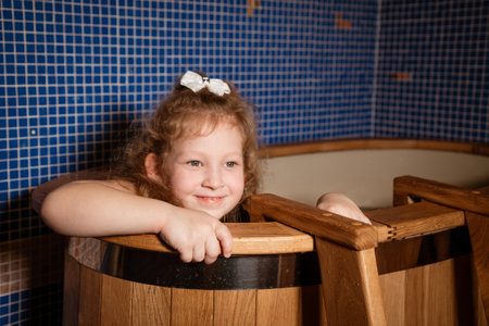 A young girl is sitting in bathtub with a wooden seat. She is smiling and looking at camera