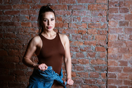 A woman in a brown tank top is standing in front of brick wall. She is wearing a denim jacket and...