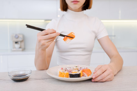 woman is eating sushi with chopsticks. plate is full sushi, and there is a small bowl soy sauce on the table. The woman is focused on her meal, and the atmosphere seems calm and relaxedの写真素材