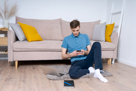A man is sitting on the floor in front of a couch, using his cell phone. The room is decorated with yellow pillows and a laptop. The man is focused on his phoneの写真素材
