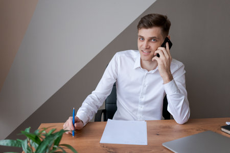 A man is talking on his cell phone while sitting at a desk with a pen and a piece of paper