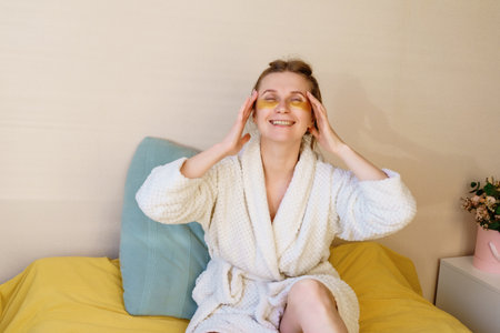 A woman is sitting on a bed with a yellow blanket and a white robe. She is wearing a yellow eye mask and has a yellow eye liner. She is smiling and she is enjoying her timeの写真素材