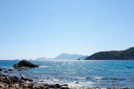 Coastal scene with a rocky shore in the foreground and mountains in the background under a clear blue sky. Concept of tranquility and nature's beautyの写真素材
