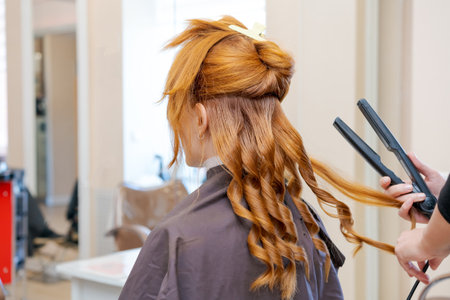 A woman with red hair is getting her hair curled by a stylist. The stylist is holding a flat iron and is working on the woman's hairの写真素材