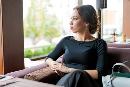 A woman is sitting on a couch with her hand on her hip. She is wearing a black dress and has a handbag on her lapの写真素材