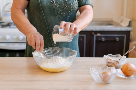 Woman is making a cake and adding flour to a bowl. The kitchen is well-equipped with a stove, oven, and sinkの写真素材