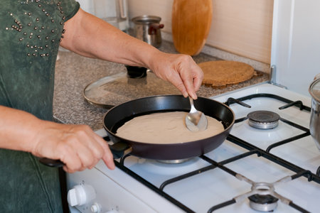 Woman is cooking a pancake on a stove. She is using a spoon to scoop the pancake batter into the panの写真素材