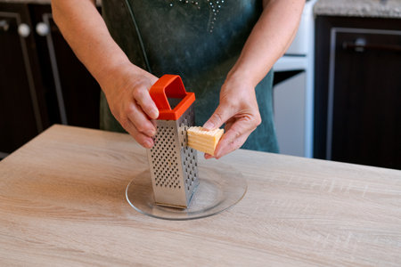 Woman is using a cheese grater to grate cheese. The cheese is on a plate and the grater is on a...