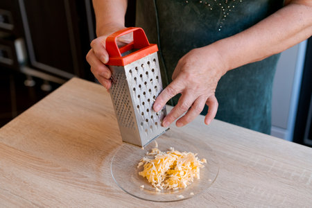 Woman is using a cheese grater on a wooden table. The cheese is being gratified into a plateの写真素材