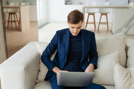 Man in a suit is sitting on a couch and using a laptop. He is wearing a blue jacket and a black shirtの写真素材