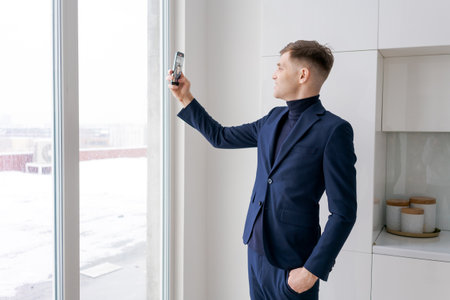 Man in a suit is taking a picture of himself with a cell phone. He is standing in front of a window