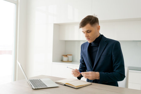 Man in a suit is sitting at a table with a laptop and a notebook. He is writing in the notebookの写真素材