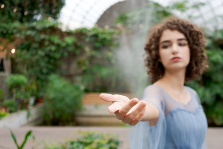 Woman in a blue dress is standing in a greenhouse. She is holding her hand out in the air