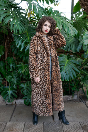 Woman wearing a leopard print coat poses in front of a green plant. She is wearing black boots and a...